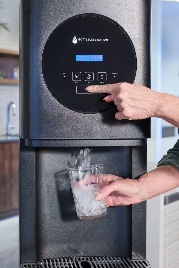 A countertop water dispenser with a black circular control panel and blue display, shown with a person's hands pressing buttons and holding a clear cup under the dispensing area.